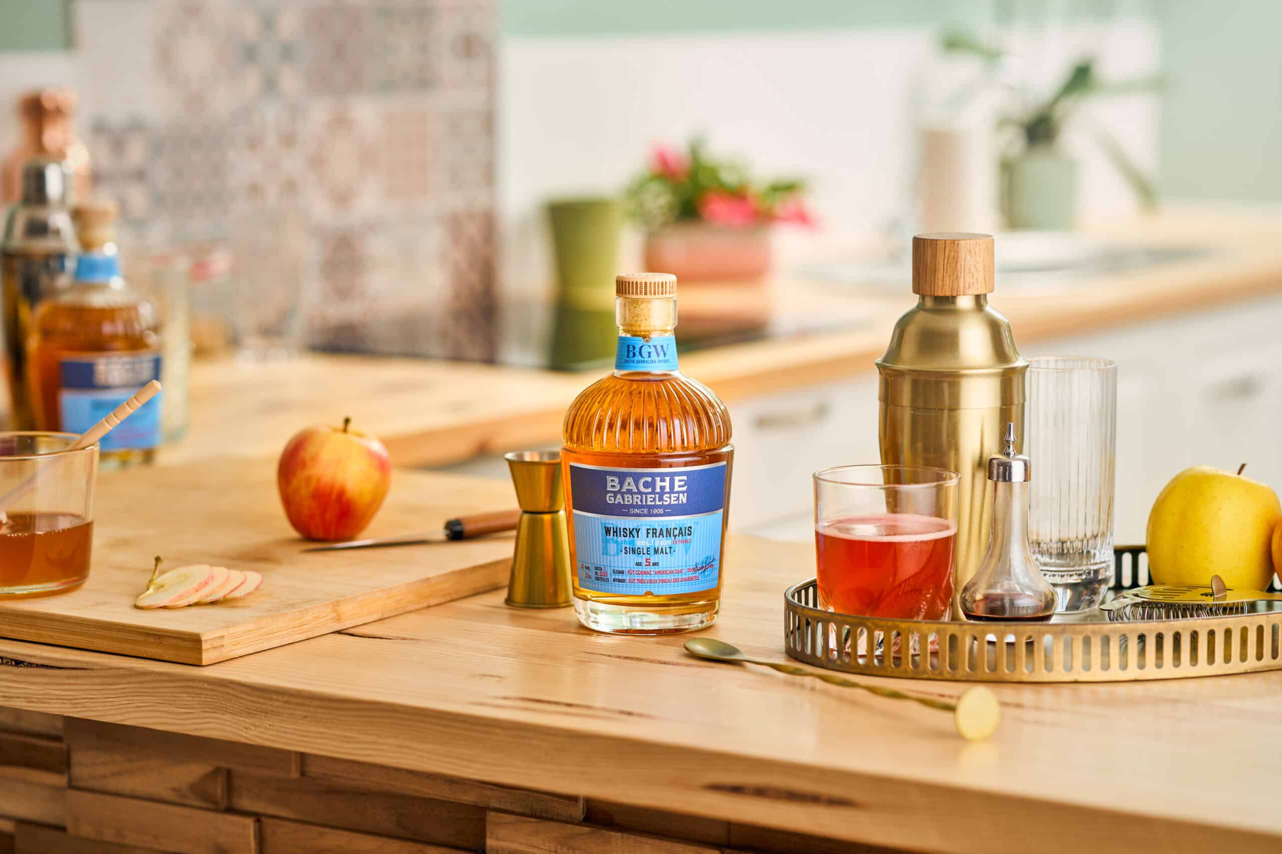 Photo du Whisky français Bache-Gabrielsen posé sur une table de cuisine avec une pomme, du miel et un shaker